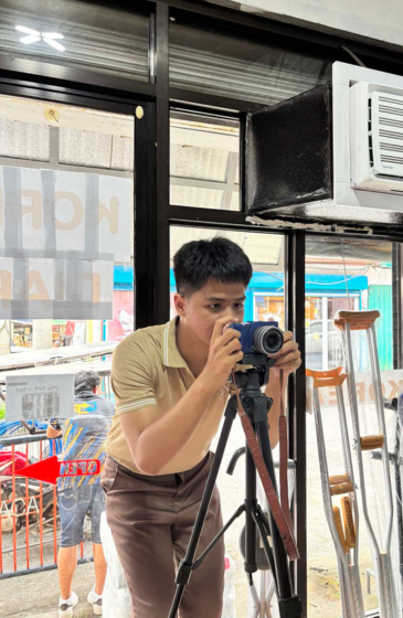 A man wearing a yellow shirt uses a camera on a tripod indoors.