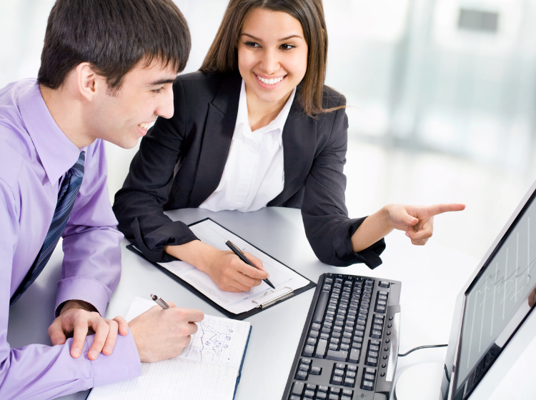 Two professionals sit at a desk, one taking notes and the other pointing at a computer monitor.