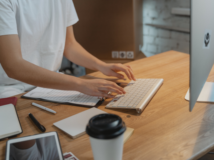 Person in a white t-shirt typing on a wireless keyboard at a wooden desk.